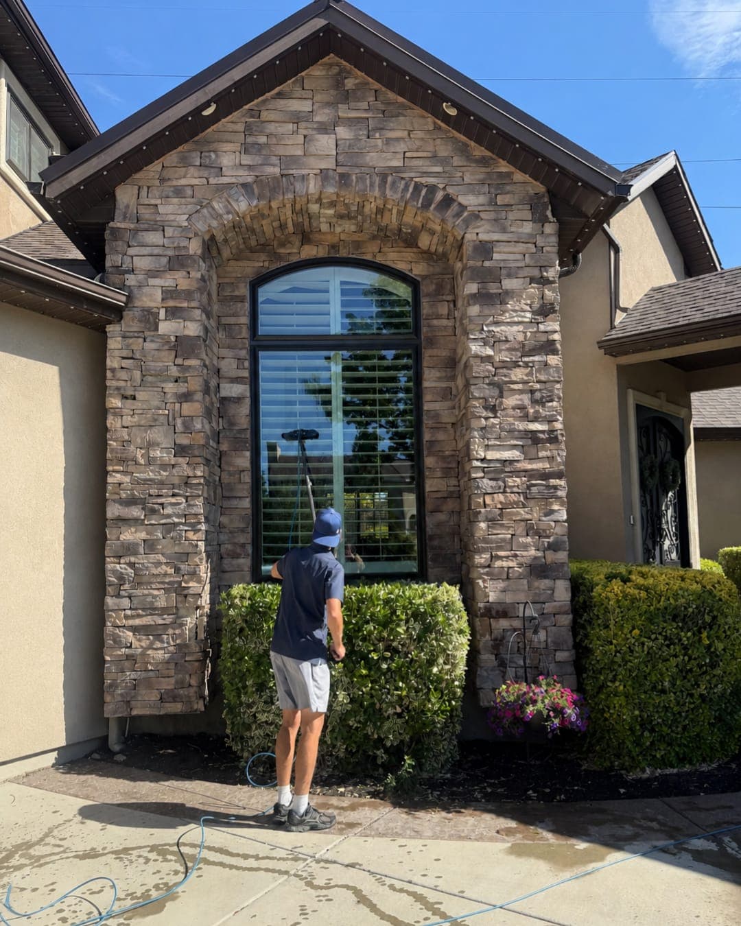 Glide technician cleaning a home's exterior window, with the roofline and gutters visible above the stone entryway